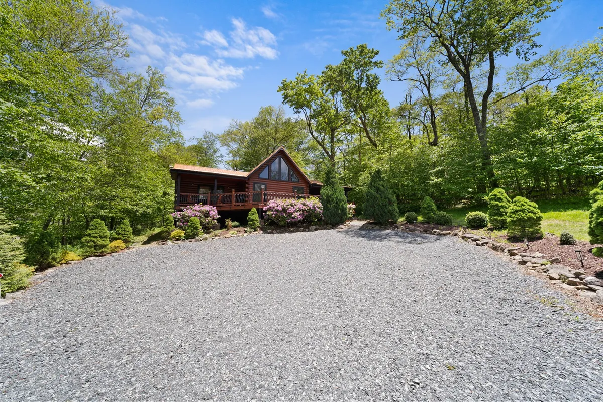 A tranquil view of the cabin exterior with flowering shrubs, gravel driveway, and expansive natural surroundings. The scene evokes a sense of peace and provides context for outdoor gatherings or quiet strolls with pets.