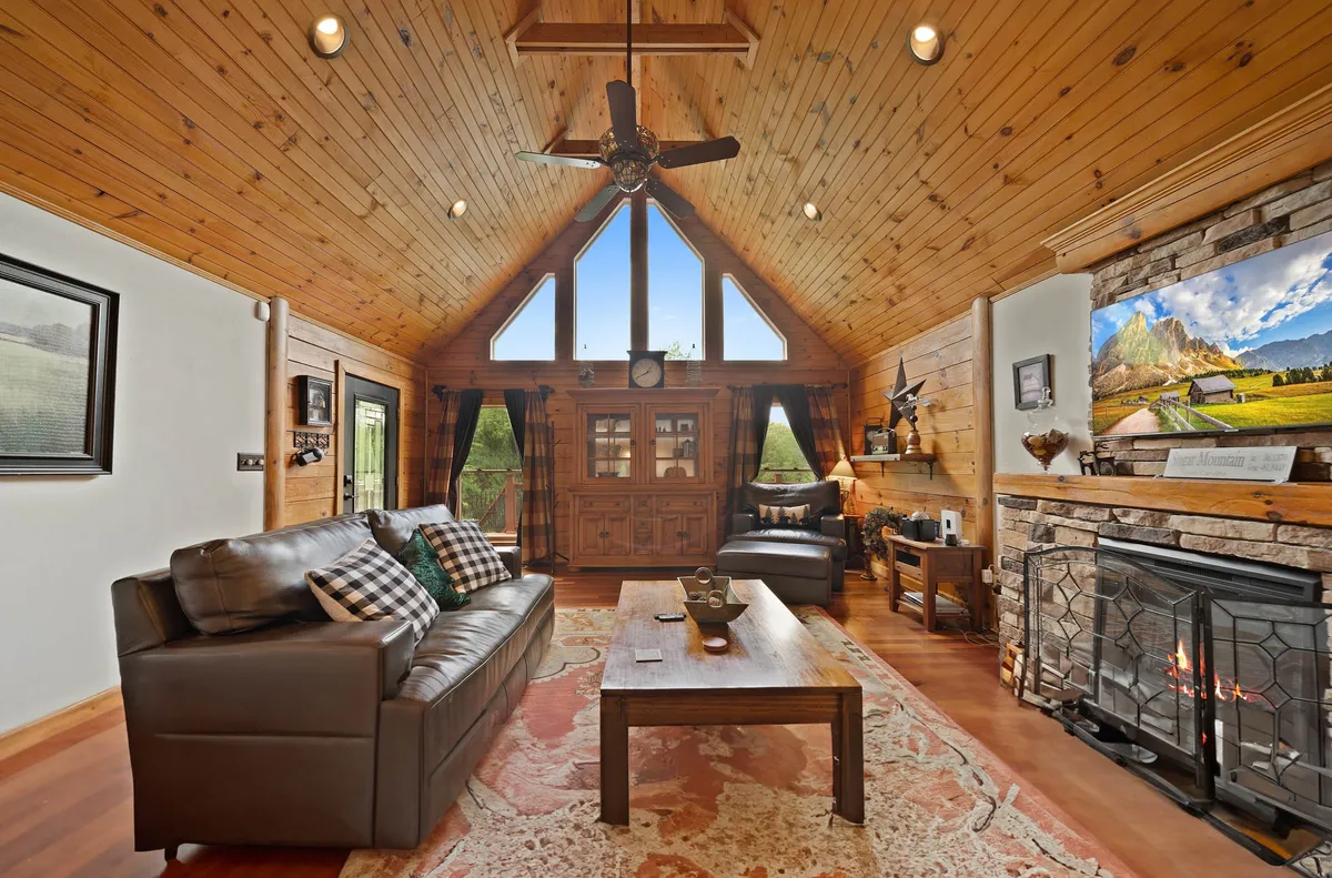 This inviting living area features expansive windows with sunrise views, a vaulted wooden ceiling, a striking stone fireplace, plush sofas, and decorative wall art. The scene speaks to comfort, warmth, and togetherness, with ample space for families to relax, watch movies, or gather by the fire after a day outdoors.