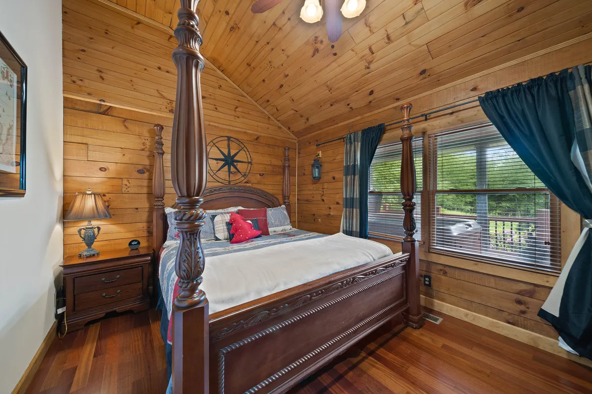 A tranquil bedroom with a four-poster bed adorned in soft bedding, wooden wall paneling, a nightstand with a lamp, and windows with curtains that allow natural light to filter in. The decor is warm, inviting, and hints at classic cabin style, creating a peaceful retreat perfect for restful sleep.