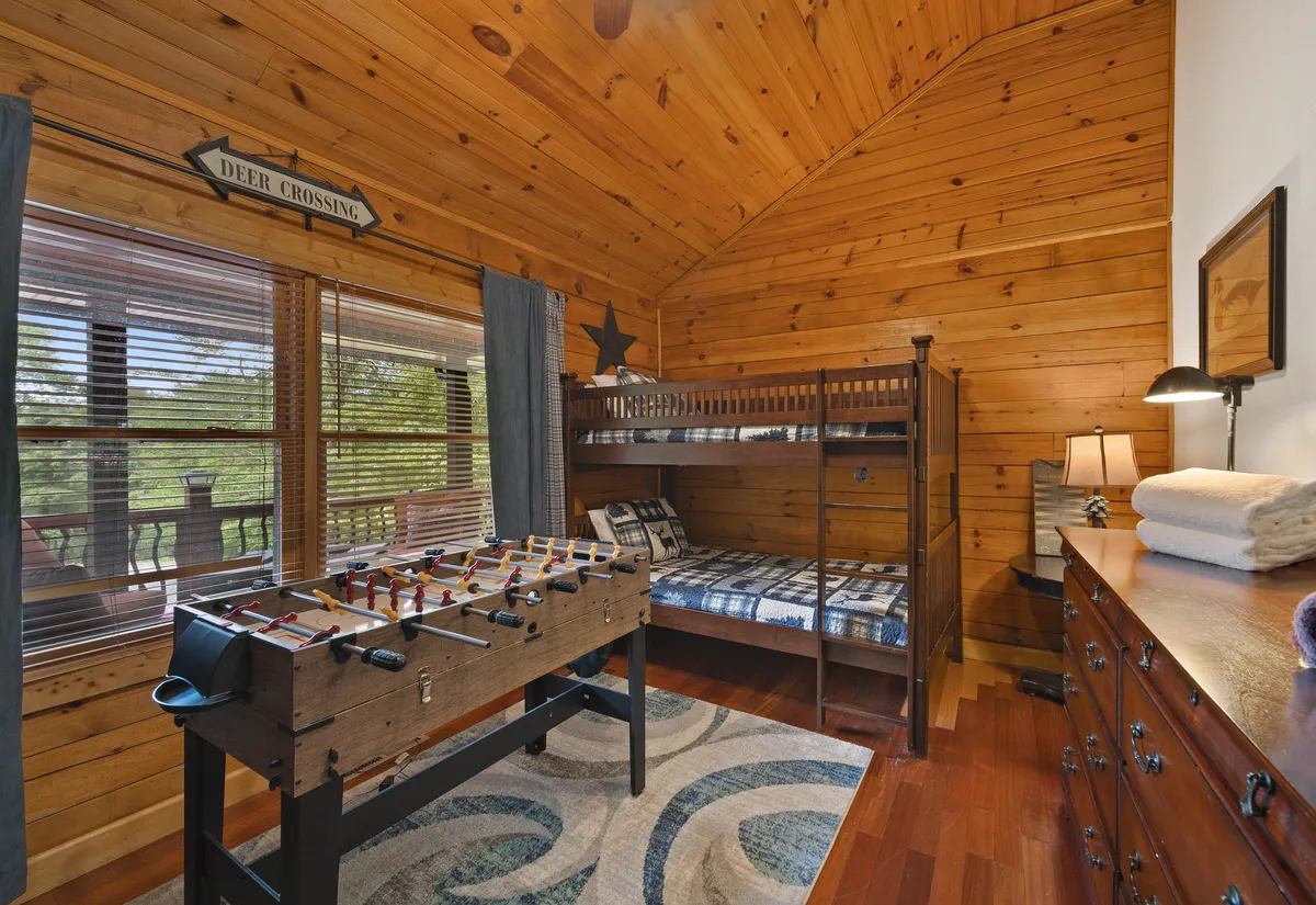 A bunk room featuring a foosball table, bunk beds, a wooden dresser, and a window with sunlight streaming in. The playful, warm vibe and family-friendly setup are ideal for both rest and fun, echoing the value of shared entertainment and togetherness.