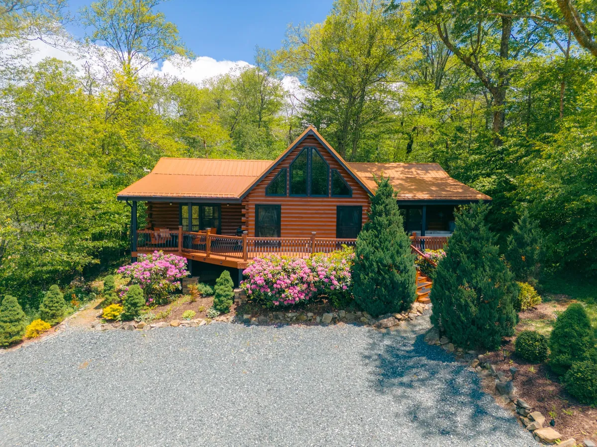 This image shows the log cabin surrounded by lush trees, flowering shrubs, and a gravel driveway. The inviting porch and natural landscaping set the scene for relaxing outdoor activities with pets. The surrounding woods are visible, hinting at trails and nature right outside the doorstep.
