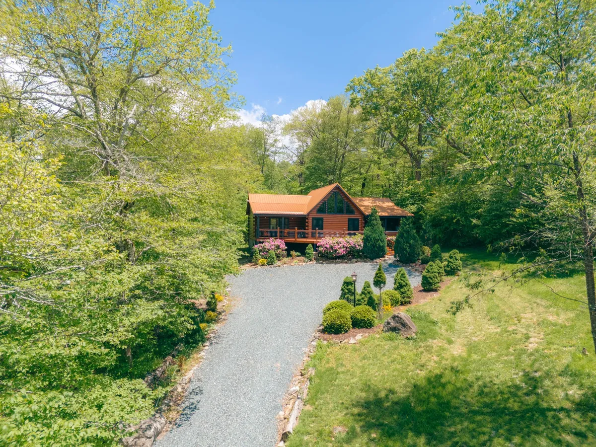 This exterior image showcases the log cabin set amidst lush trees, flower beds, and a gravel driveway. The natural surroundings emphasize the peaceful mountain setting, inviting guests to imagine outdoor activities or serene winter landscapes.