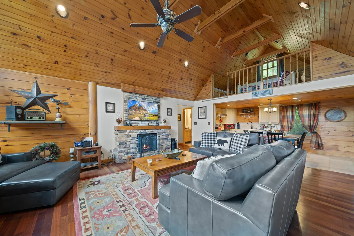 A beautifully appointed living room highlighting a glowing fireplace, comfortable sofa, and expansive windows that look out onto natural woodland. Wooden beams and a plush rug add to the inviting, romantic ambiance, making this an ideal spot for cozy evenings together.