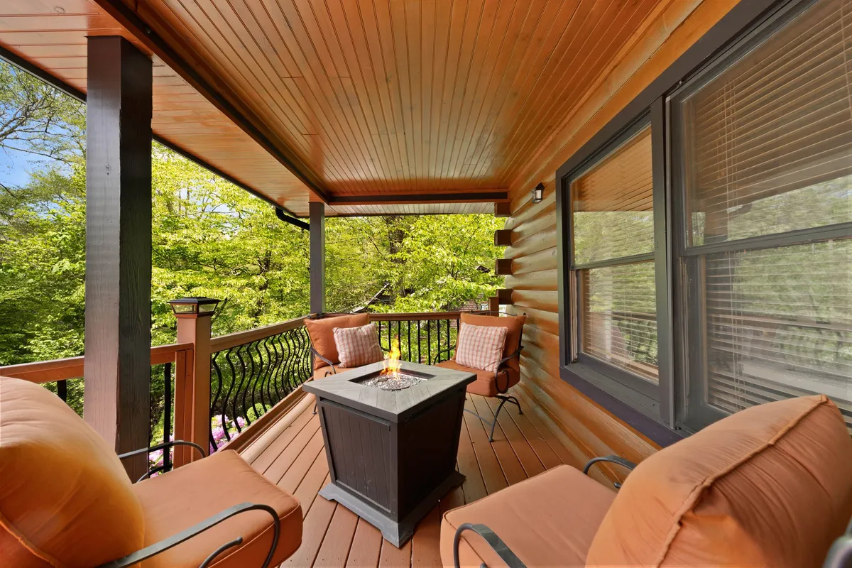 An inviting outdoor seating area on a covered deck, centered around a gas fire table. Comfortable chairs are arranged for conversation or relaxation, with forest greenery enveloping the space. The deck is clearly designed for communal gatherings, with a sense of warmth and connection to nature.