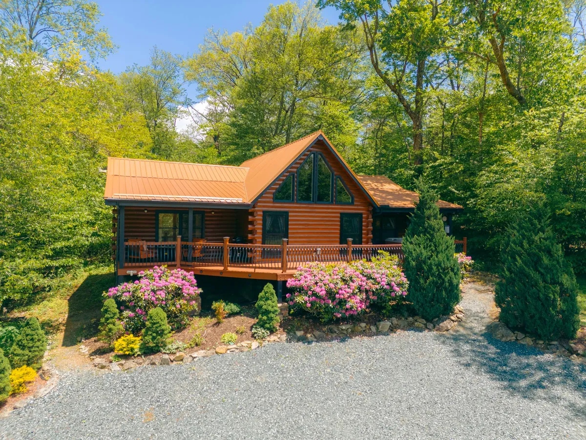 The image shows a cozy log cabin nestled among lush greenery, with vibrant flower beds and a welcoming wooden porch that invites relaxation. The setting is serene, giving the impression of seclusion and peace, perfect for outdoor gatherings or simply enjoying the mountain air.