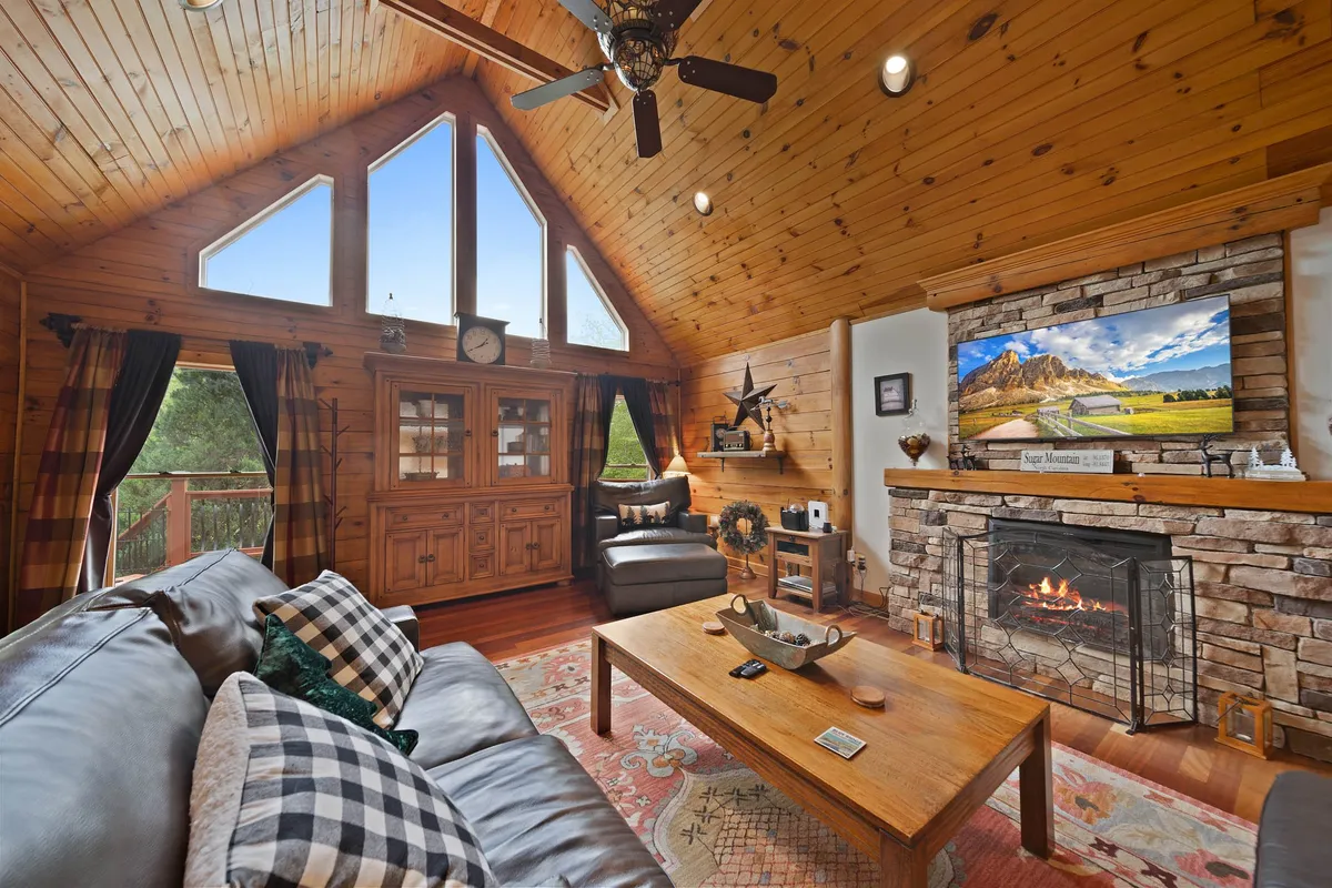 This image showcases a spacious living room featuring large windows that frame serene woodland views, a cozy gas fireplace, vaulted wooden ceilings, comfortable seating, and inviting decorative accents. The warmth of the space and the expansive windows highlight the seamless connection to nature mentioned in the section.