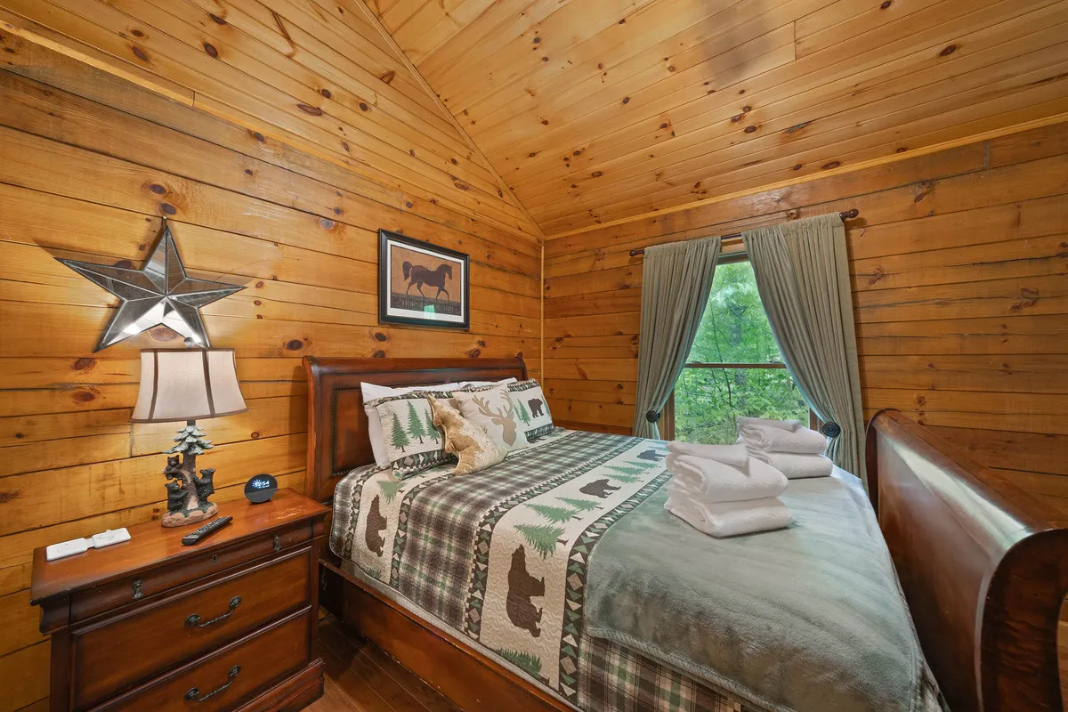 A rustic bedroom featuring wooden walls, a bed dressed with decorative bedding, a star ornament, a bedside lamp, and a curtained window. The view outside offers a glimpse of the surrounding nature, enhancing the room's peaceful ambiance.