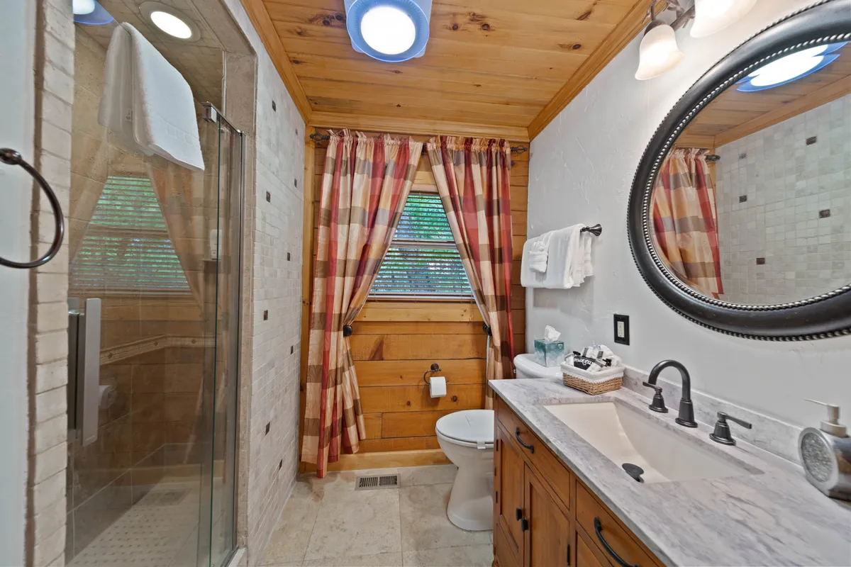 A well-appointed bathroom with wooden cabinetry, a large mirror, plaid curtains, and a shower area. The space feels homey and serene, designed for comfort and practical use with charming details that echo the cabin's style.