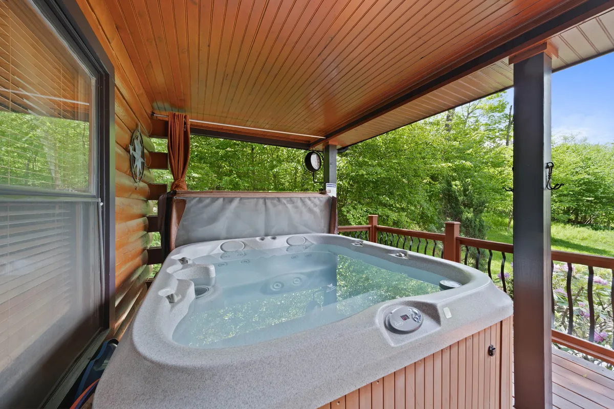 A tranquil scene of a wooden deck nestled among lush greenery, featuring a bubbling hot tub as the central element. The hot tub is surrounded by ample deck space and offers sweeping views of the forest, hinting at the seclusion and serenity of the setting. The image captures golden hour lighting, evoking the experience of relaxing during sunset or stargazing at night.