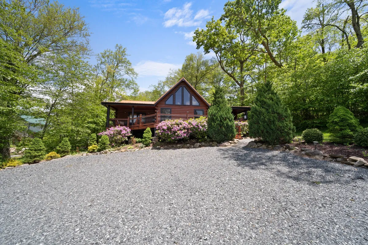 This exterior shot captures the log cabin nestled among flowering shrubs, with a gravel driveway and lush greenery surrounding the home. The inviting porch suggests a space perfect for relaxing after a day of adventure, while the bloom-filled landscape hints at the abundant nearby natural beauty and exploration opportunities.