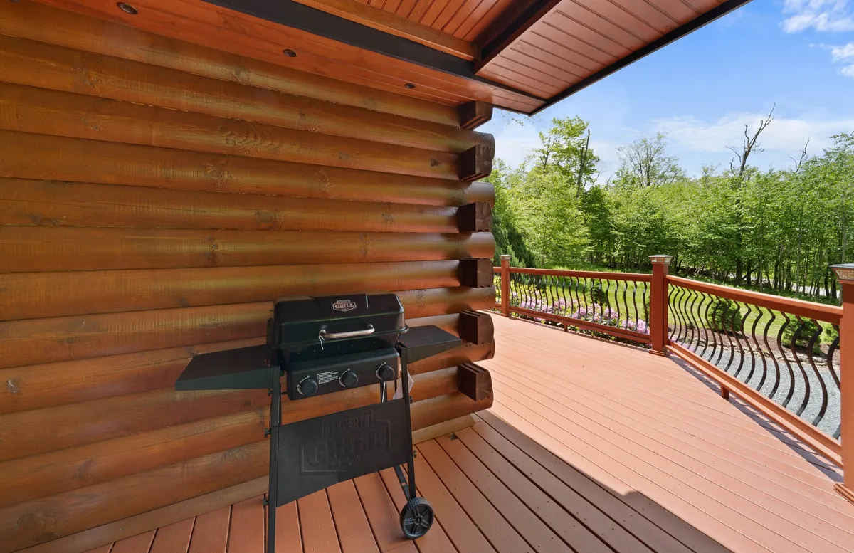 A close-up exterior view of the cabin deck, featuring a grill, log wall, and sturdy deck railing. The setup is clearly designed for barbecues and relaxed outdoor gatherings, with the surrounding trees providing a sense of immersion in nature.